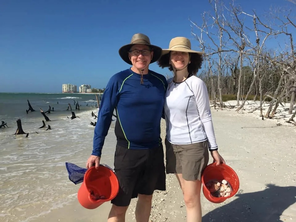 Two guests in sun hats hold red buckets of shells on a driftwood beach