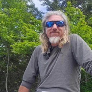 Capt. Shawn Palmer with long hair and sunglasses in front of green trees