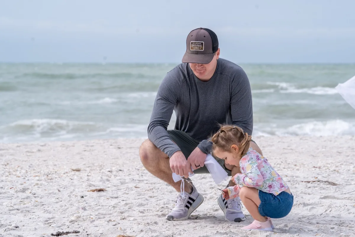 A man kneels beside a toddler on the beach as she looks for shells