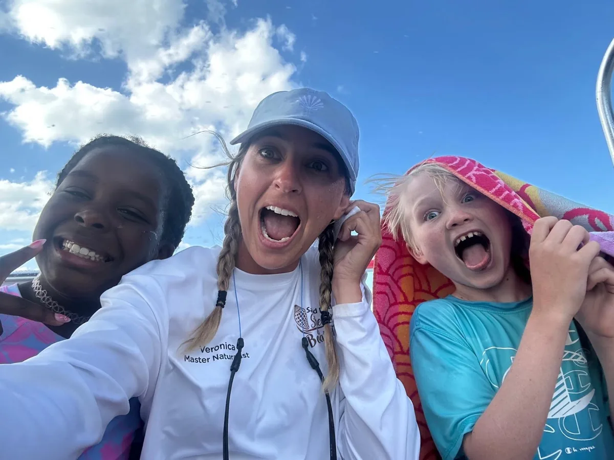 A crew member in a white shirt takes a selfie on the boat with two laughing kids